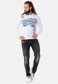 Weißes Langarmhemd mit blauem Grafikdesign und Spritzakzenten, kombiniert mit dunkelblauen Jeans mit strukturierten Details und schlanker Passform. Weiße Sneaker.