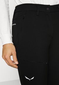 Calças de yoga pretas com um bolso com fecho, fecho de botão e um detalhe de logótipo branco. O tecido tem uma aparência texturizada.