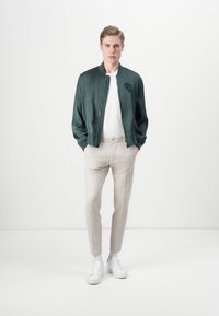 Green bomber jacket with a front zipper, white T-shirt, beige trousers, and white sneakers. Smooth texture and subtle logo detail on the jacket.