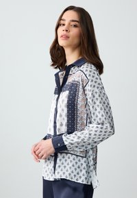 Chemisier à manches longues en tissu léger, présentant un mélange de motifs en bleu marine et blanc, avec un col classique et une fermeture à boutons.