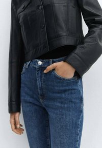 Veste courte en cuir noir avec fermeture à boutons, associée à un jean en denim bleu. Le jean présente un bouton contrastant et des détails de poche.