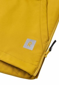 Keltainen takki, jossa on sileä pinta, hopeinen suorakulmainen logo ja musta vetoketju. Yksinkertainen muotoilu siisteillä ompeleilla.