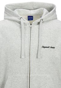Haut de hoodie zippé couleur gris clair en tissu doux, doté d'une capuche, de cordons de serrage et de poches avant. "Original Studio" est brodé en noir.