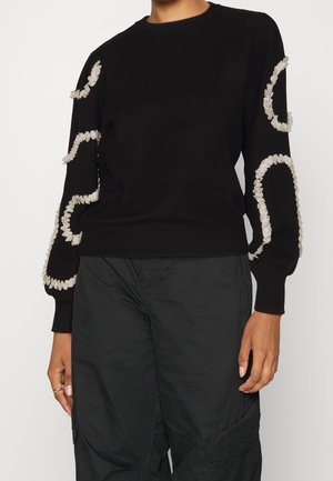 Pull noir à manches longues avec des motifs circulaires blancs froncés sur les manches, porté avec un pantalon noir, présenté de la nuque jusqu'à mi-cuisse.