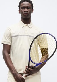 Μπεζ πόλο πουκάμισο με διακριτικό ριγέ σχέδιο, με κοντά μανίκια και γιακά. Το μοντέλο κρατάει ρακέτα τένις με μπλε πλαίσιο και κίτρινα χορδές.