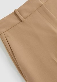 Beežid püksid, millel on siledat tekstuur, lame esikülg ja vööpaelad. Kanga pinnal on peen diagonaalne kudumismuster.