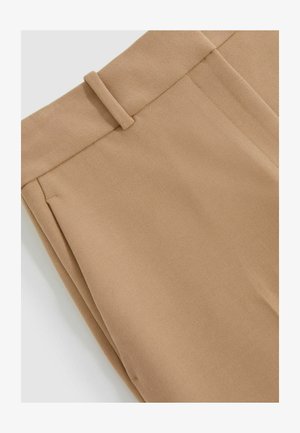 Beežid püksid, millel on siledat tekstuur, lame esikülg ja vööpaelad. Kanga pinnal on peen diagonaalne kudumismuster.