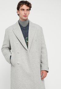 Dvojradový šedý kabát s houndstooth vzorom s veľkými gombíkmi a bočnými vreckami, položený na textúrovanej patterned roláku.