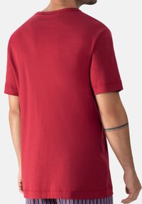 Man in een eenvoudige rode T-shirt met korte mouwen gezien vanaf de achterkant, met een zwart dubbelbandtattoo op zijn linkerarm.