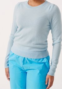 Persoon draagt een lichtblauwe trui met lange mouwen en felblauwe broek, staand tegen een effen witte achtergrond.