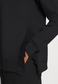 Svart sweatshirt med ribbad nederkant och sidodetaljer med volanger. Mjukt tyg och åtsittande muddar fulländar designen.