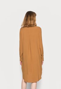 Robe à manches longues dans une teinte marron chaude, fabriquée en tissu doux et fluide, avec un pli central et des poignets boutonnés, dotée d'un ourlet incurvé.