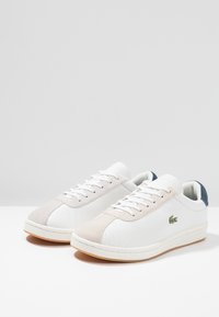 Baskets en cuir blanc avec des accents en suede gris clair, bout rond, semelle en caoutchouc plate et logo vert sur le côté. Lacets et col rembourré.