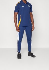 Homme portant un polo bleu Real Madrid avec des accents jaunes, un pantalon de survêtement bleu assorti avec des rayures blanches, et des chaussures de sport noires.
