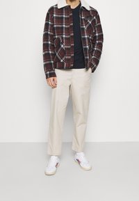 Geruite jas in donkerrood, wit en zwart met een witte bontkraag; eronder een zwart T-shirt, crèmekleurige broek en witte sneakers met gekleurde strepen.