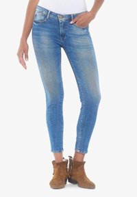 Kapelevat siniset farkut, joissa on haalistunut pesu, viisi taskua ja vetoketju. Yhdistettynä ruskeisiin nilkkureihin.