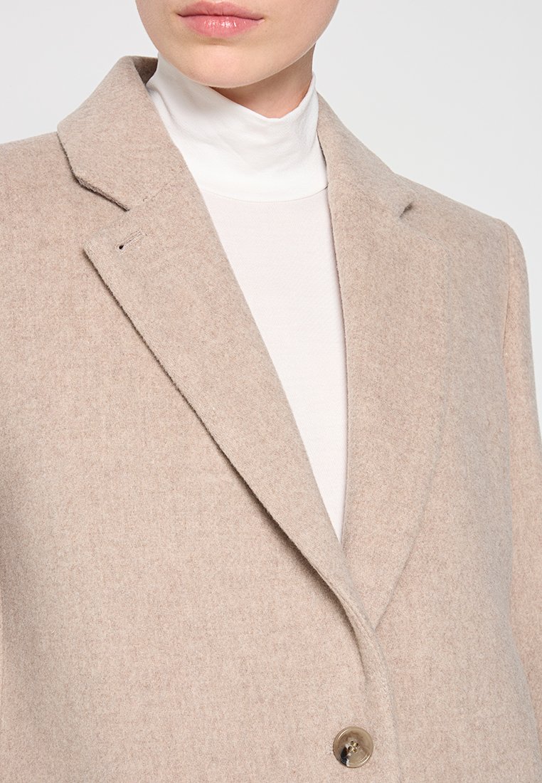 Beige Wollmischung-Blazer mit Reverskragen, Ein-Knopf-Verschluss, strukturiertem Schnitt und glatter Textur, getragen über einem weißen Rollkragenpullover.