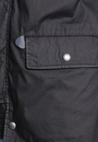 Veste en tissu noir dotée d'une poche à rabat fermée par un bouton-pression métallique et d'une ouverture latérale. Le matériau présente une finition lisse avec une légère texture.