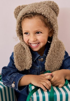 Beige, flauschige Bärenmütze mit Ohrenakzenten, kombiniert mit einer Jeansjacke. Im Vordergrund befinden sich grün-weiß gestreifte Geschenkeboxen.