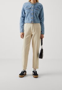 Vrouw in een lichtblauwe denimjack, beige broek, zwart-witte sneakers, die een kleine zwarte handtas met een polsbandje vasthoudt.
