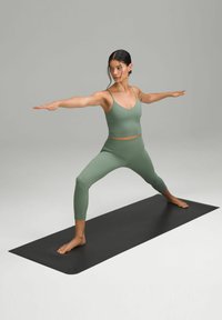 Zelený jóga outfit s přiléhavým topem a legínami s vysokým pasem, pózující na černé jógové podložce, zdůrazňující hladkou látku a štíhlý design.