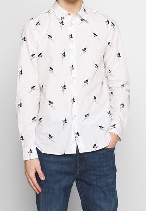 Mand iført hvid skjorte med knapper med sort-hvide tegneserieansigtsmønstre og blå jeans, stående med den ene hånd i lommen.