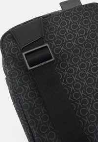 Borsa nera con motivo, caratterizzata da una texture liscia, cinturino in nylon regolabile e dettaglio del logo in rilievo. Forma rettangolare compatta con un design elegante.