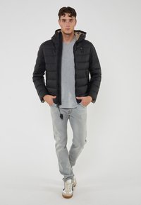 Joven de pie vestido con una chaqueta acolchada con capucha negra, jeans gris claro, camiseta gris y zapatillas blancas con suelas marrones sobre un fondo blanco.