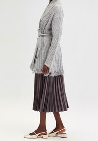 Šedý žebrovaný kardigan s třásněmi na lemu, zavázaný v pase. Kombinován s plisovanou sukní v délce ke kolenům v tmavě šedé a bílými botami na robustním podpatku.