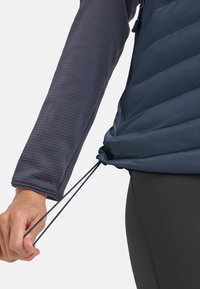 Blauer isolierter Weste mit einer geschnürten Kordel, über einem strukturierten dunkelgrauen Langarmshirt. Glattes Gewebe mit puffigen Einsätzen.