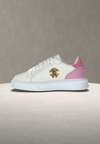 Λευκό χαμηλό αθλητικό παπούτσι με ροζ διακόσμηση στη φτέρνα και χρυσό διακοσμητικό λογότυπο στο πλάι, τοποθετημένο σε μπεζ επιφάνεια με ουδέτερο φόντο.