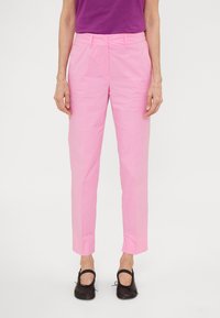 Persoon in lichtroze rechte broek, zwarte Mary Jane schoenen en een paars shirt in de broek, staande tegen een witte achtergrond.