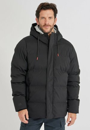 Mann trägt eine schwarze Steppjacke mit Kapuze, roten Kordelenden und dunkle Cargohosen, steht vor einem einfarbigen Hintergrund.