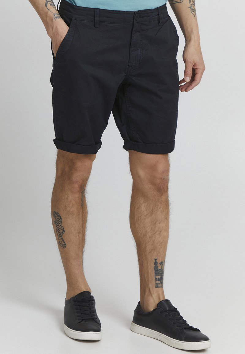 Shorts en coton noir avec des revers, dotés de poches latérales et d'un design à l'avant plat. Portés avec des baskets noires sur une peau texturée.