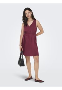 Robe bordeaux avec un décolleté en V et une taille élastique, associée à un sac à main noir et des ballerines à pois. Tissu doux et design mi-long.