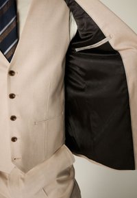 Veste de costume beige ouverte montrant une doublure noire à motifs, associée à un gilet beige et une cravate rayée bleue, marron et blanche.