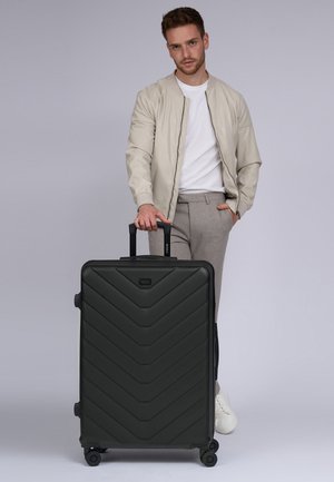 Schwarzer Koffer mit Zickzackmuster, aus hartem Material, mit einem ausziehbaren Griff und vier Rädern. Ein Mann in legerer Kleidung steht daneben.