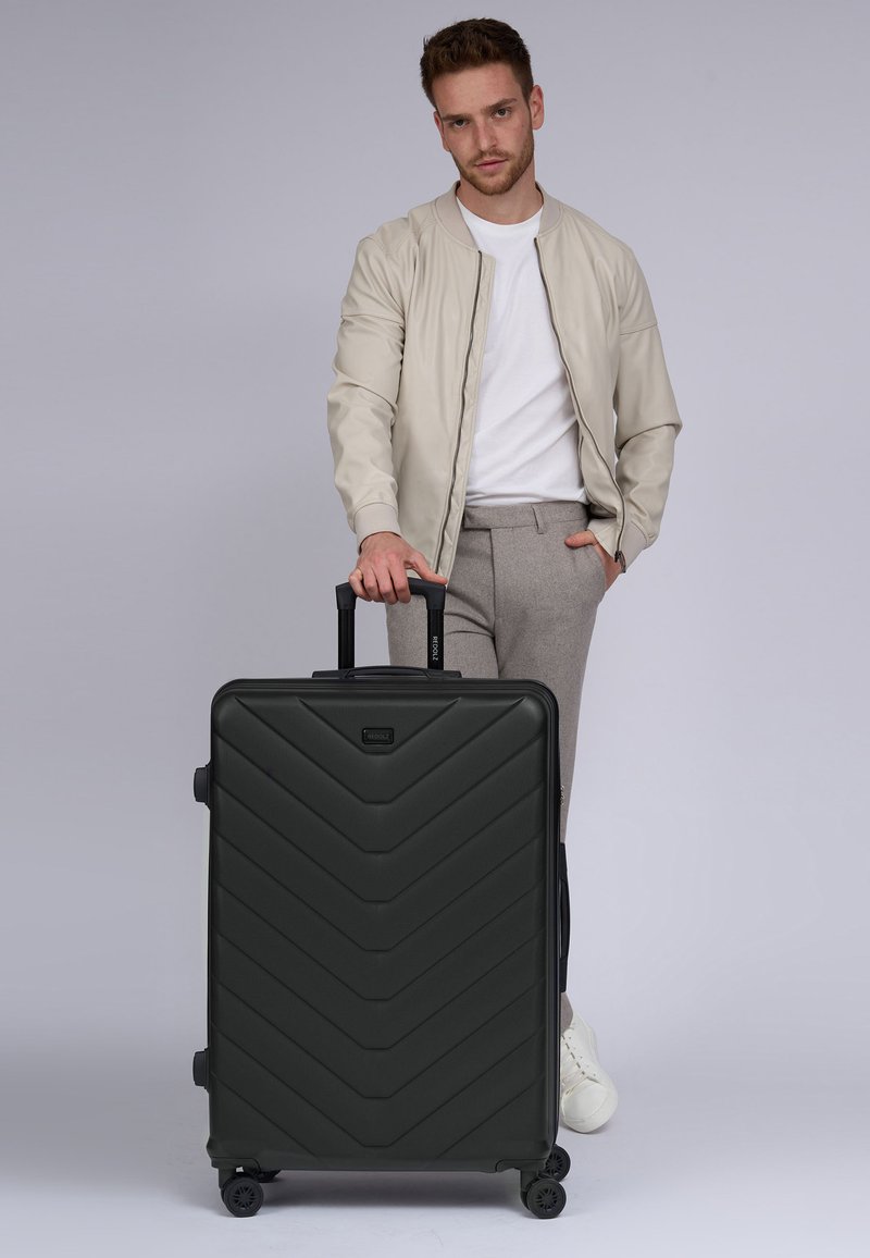 Valigia nera con un motivo a zigzag, materiale rigido, manico estraibile e quattro ruote. Un uomo in abbigliamento casual è in piedi accanto ad essa.