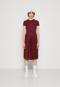 Donna con un cappello bianco, una maglietta attillata bordeaux, una gonna midi plissettata, calze bianche e sneakers bianche, in piedi su uno sfondo neutro.