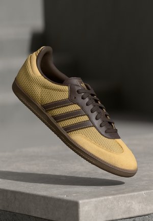 Gul og brun sneaker med tekstureret stof og tre striber, ophængt over en grå betonforkant mod en mørk sløret baggrund.