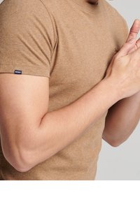 Man som bär en brun t-shirt med kort ärm, med händerna knäppta nära bröstet mot en enfärgad ljus bakgrund.