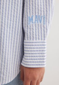 Blue and white striped shirt featuring an embroidered logo "MAVI" on the cuff, made of lightweight fabric with a textured finish.