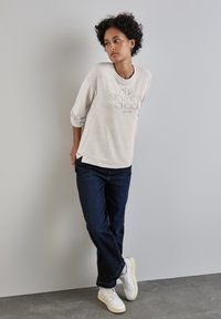 Pullover grigio chiaro con testo, polsini a costine e spacchi laterali. Indossato con jeans larghi blu scuro e sneakers bianche con suole testurizzate.