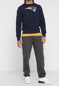 Sudadera azul marina con el logotipo de la NFL, combinada con pantalones de chándal grises. Zapatillas deportivas blancas completan el atuendo. Camiseta amarilla visible debajo.
