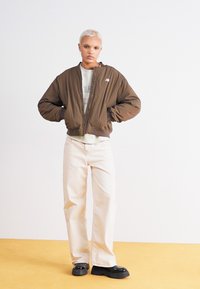 Pessoa com cabelo loiro curto, vestindo uma jaqueta bomber marrom, um sweatshirt cinza claro, calças largas de cor creme e sapatos pretos de sola grossa, em pé dentro de casa.