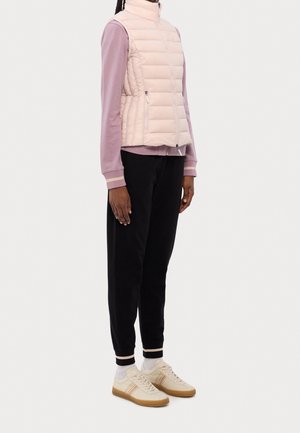 Eine Person trägt eine hellrosa Steppweste über einem mauvefarbenen Zip-Up-Oberteil, schwarze Jogginghosen mit gerafften Knöcheln, weiße Socken und beigefarbene Turnschuhe.