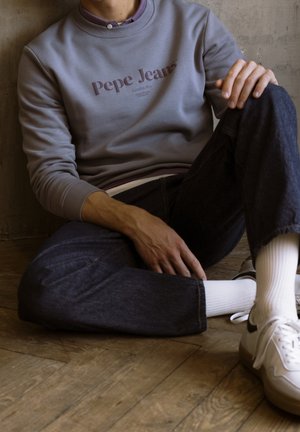 Šedá mikina s logem "Pepe Jeans", tmavé džíny, bílé sportovní boty a bílé ponožky. Materiál vypadá měkce a design je ležérní.