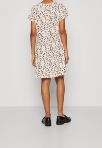 Vestido de manga corta en un tejido crema con un patrón floral en tonos rojos y negros. Diseño de corte holgado, hasta la rodilla. Zapatos negros complementan el conjunto.
