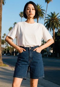 Tânără stând afară cu mâinile în buzunare, purtând un tricou alb și pantaloni scurți din denim cu talie înaltă, cu palmieri vizibili în fundal.