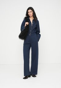 Πουκάμισο από denim σε σκούρο μπλε με κούμπωμα κουμπιών, σετάρισμένο με μπλε σκούρο παντελόνι φαρδιάς γραμμής με ζώνη, συμπληρωμένο από μια μαύρη τσάντα με απαλό, χνουδωτό ύφασμα.